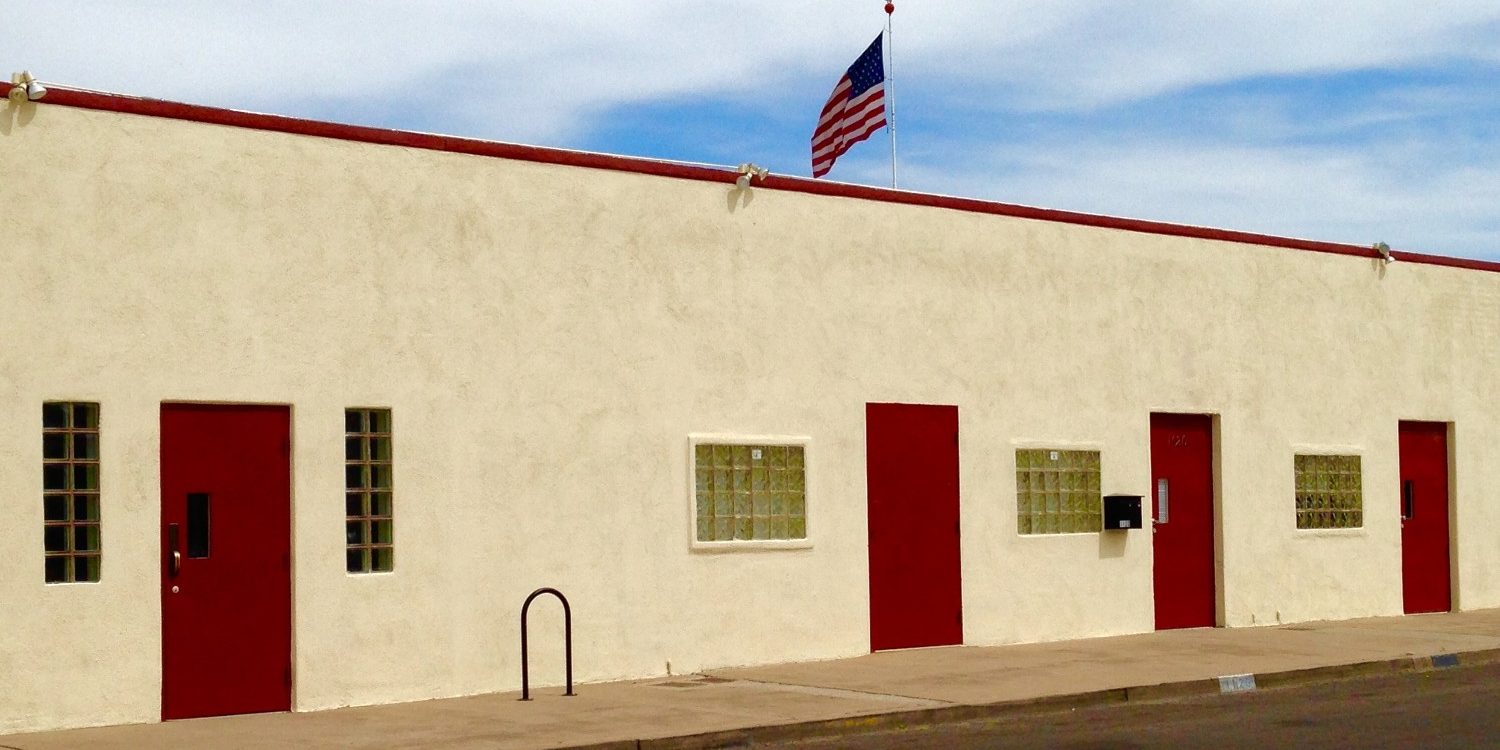 Professional commercial exterior painting in Jacksonville Beach, featuring a low-rise commercial building with fresh beige and maroon stucco paint, glass block windows, red doors, and a flying American flag against a cloudy sky.
