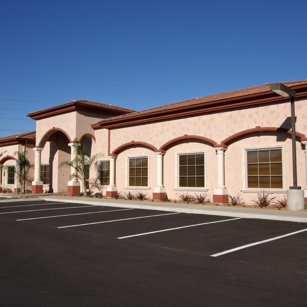 Professional commercial exterior painting in Jacksonville Beach, featuring a low-rise commercial building with fresh beige and maroon stucco paint, arched windows, and a paved parking lot under a clear blue sky.